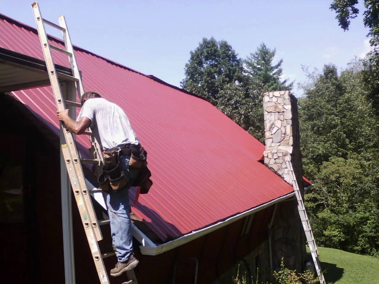 Roofer on a ladder beside a red metal roof and stone chimney — Soffit & Fascia Repair by Roofing Brothers in Thurston